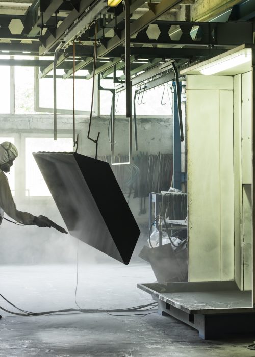 view of a worker wearing a full white protective suit and breathing mask, sand blasting a metal crate hung from a metal beam in the ceiling of an industrial hall